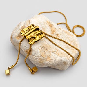 Gold bolo tie lariat necklace arranged over a textured white stone on a clean neutral background. The polished sculptural wave slider sits above two long tassel chains with geometric square charms, while the continuous snake chain curves around the stone. Product photography highlights the organic statement design, rich gold finish and adjustable long Y necklace construction.