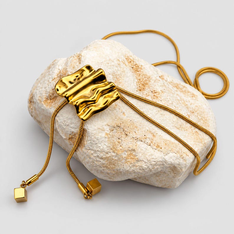 Gold bolo tie lariat necklace arranged over a textured white stone on a clean neutral background. The polished sculptural wave slider sits above two long tassel chains with geometric square charms, while the continuous snake chain curves around the stone. Product photography highlights the organic statement design, rich gold finish and adjustable long Y necklace construction.