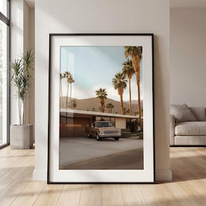 May include: Framed print featuring a mid-century modern home with a brown pickup truck parked in the driveway. Palm trees line the street, and mountains are visible in the background. The print is displayed in a room with hardwood floors and a large window.