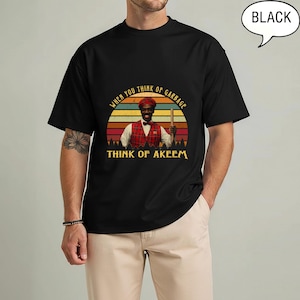 May include: Black t-shirt featuring a graphic of a man in a red plaid vest and the text "When you think of garbage, think of Akeem." The design is set against a sunset backdrop. The shirt is a classic crew neck.