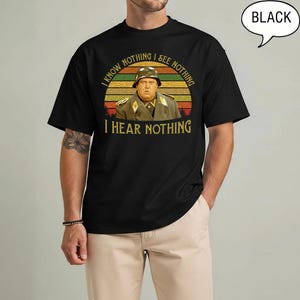 May include: Black t-shirt featuring a vintage-style graphic of a man in a military uniform with the text "I KNOW NOTHING I SEE NOTHING I HEAR NOTHING" in yellow and white. The graphic is set against a sunset-colored background.