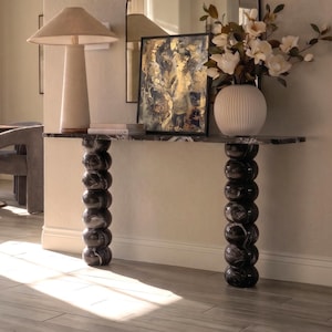 May include: A console table featuring a long, rectangular black marble top with white veining, supported by two vertical columns composed of stacked, rounded black marble spheres. The table surface holds a framed abstract painting and a white vase.