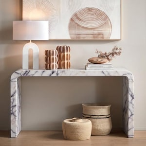 May include: A white marble console table with grey veining, featuring a white arched table lamp, two fluted wooden vases, a stack of books with a decorative bowl, and two woven baskets on the floor. A framed abstract art piece hangs above.