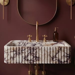 May include: A rectangular wall-mounted marble sink featuring a fluted exterior design with white and deep purple veining. The sink is equipped with a tall brass faucet, two matching brass handles, and a brown glass soap dispenser on the basin edge.