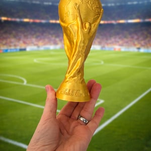 Puede incluir: Un trofeo dorado con un globo en la parte superior y una base curva. Las palabras "FIFA WORLD CUP" son visibles en la base. El trofeo se sostiene en una mano sobre un fondo borroso de un campo de fútbol y un estadio.