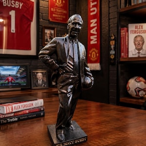 May include: A dark bronze-toned statue of Sir Matt Busby standing on a rectangular base with his name engraved in gold lettering. The figure is depicted wearing a suit and tie, with one hand resting on the hip and the other holding a football.