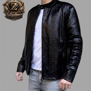 Peut inclure: Veste en cuir noire avec une texture peau de serpent, col montant et fermeture éclair. La veste est portée sur un t-shirt blanc et associée à un jean gris. Le logo Zafleatherco est visible en haut à gauche.