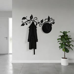 May include: A black metal coat rack with a leaf and vine design mounted on a wall. A black coat and a black hat are hanging from the rack. A potted plant is to the right. The wall is white.