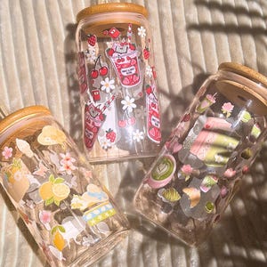 Könnte beinhalten: Drei transparente Glasbecher mit Bambusdeckeln und Strohhalmen. Jeder Becher ist mit farbenfrohen Illustrationen von Früchten, Blumen und Getränken verziert. Einer zeigt Erdbeeren und Kirschen, ein anderer Zitronen und der dritte Gebäck.