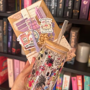 Könnte beinhalten: Ein klares Glas mit Bambusdeckel und Strohhalm, verziert mit Aufklebern von Rosen, Drachen und Büchern. Im Hintergrund befindet sich ein verpacktes Paket mit Aufklebern und dem Text "Reading Log". Das Thema ist bücherlieb und Fantasy.