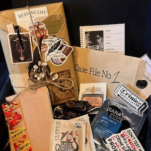 Könnte beinhalten: Eine Geschenkbox mit Krimi-Thema, die ein verpacktes Buch, Aufkleber, Teebeutel, Snacks und Hautpflegeprodukte enthält. Die Box enthält ein Lesezeichen, einen Notizblock und einen Aufkleber mit der Aufschrift "Crime Book Club".