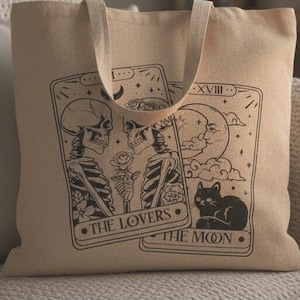 Op de afbeelding: Beige draagtas met een zwart tarotkaartontwerp met skeletten, rozen, een maan en een zwarte kat. De tekst op de tas luidt "THE LOVERS" en "THE MOON". De tas ligt op een beige bank.