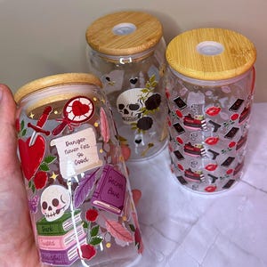 Könnte beinhalten: Drei Glasbecher mit Bambusdeckeln. Die Becher sind mit gotischen Illustrationen verziert, darunter Totenköpfe, Herzen, Rosen und der Text "Danger Never Felt So Good" und "Secret Love".