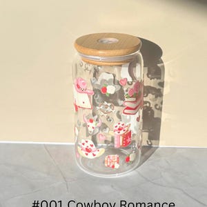May include: A clear glass tumbler with a wooden lid, decorated with pink and red cowboy-themed illustrations. The design includes hearts, books, and cowboy hats. The text "#001 Cowboy Romance" is printed at the bottom.