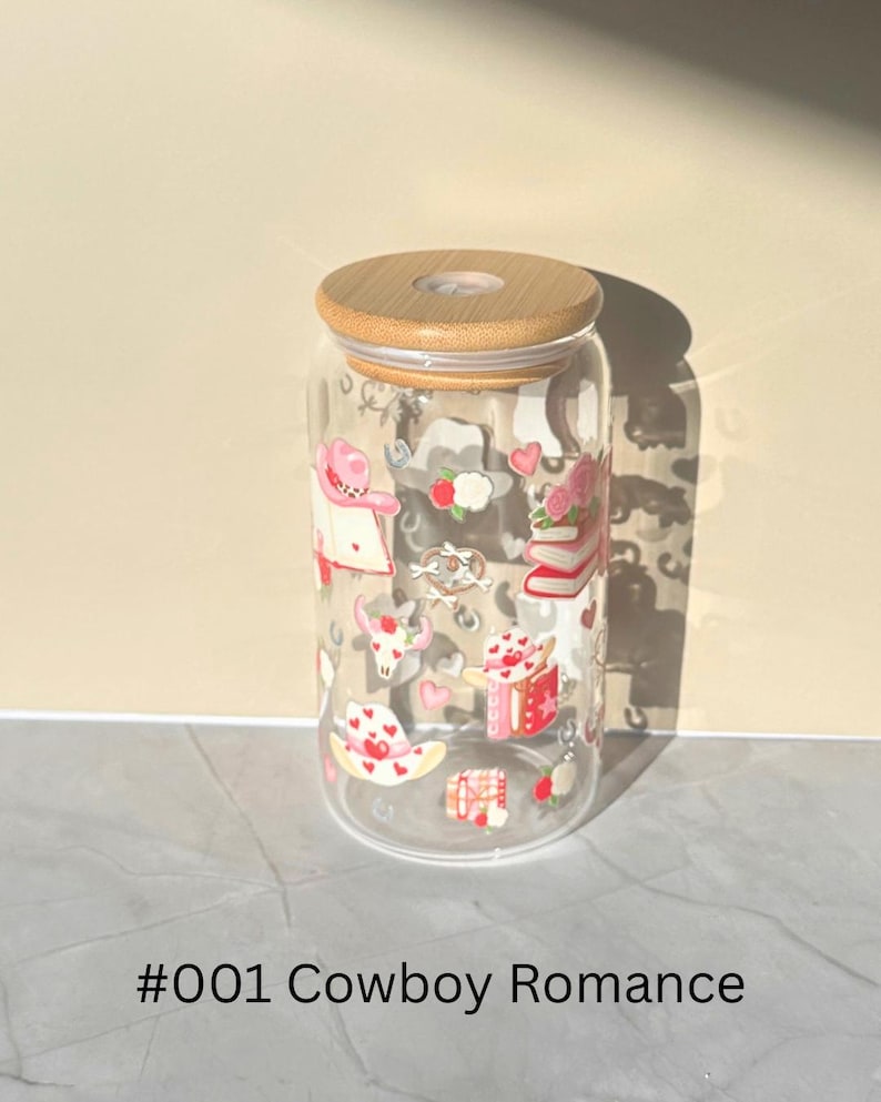 May include: A clear glass tumbler with a wooden lid, decorated with pink and red cowboy-themed illustrations. The design includes hearts, books, and cowboy hats. The text "#001 Cowboy Romance" is printed at the bottom.