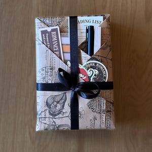 May include: A wrapped gift with a black ribbon and bow. The wrapping paper is brown with a vintage print. A pen, bookmark, and sticker are visible in a pocket on top. The bookmark reads "ROMANCE RO" and "Powered by and coffee".