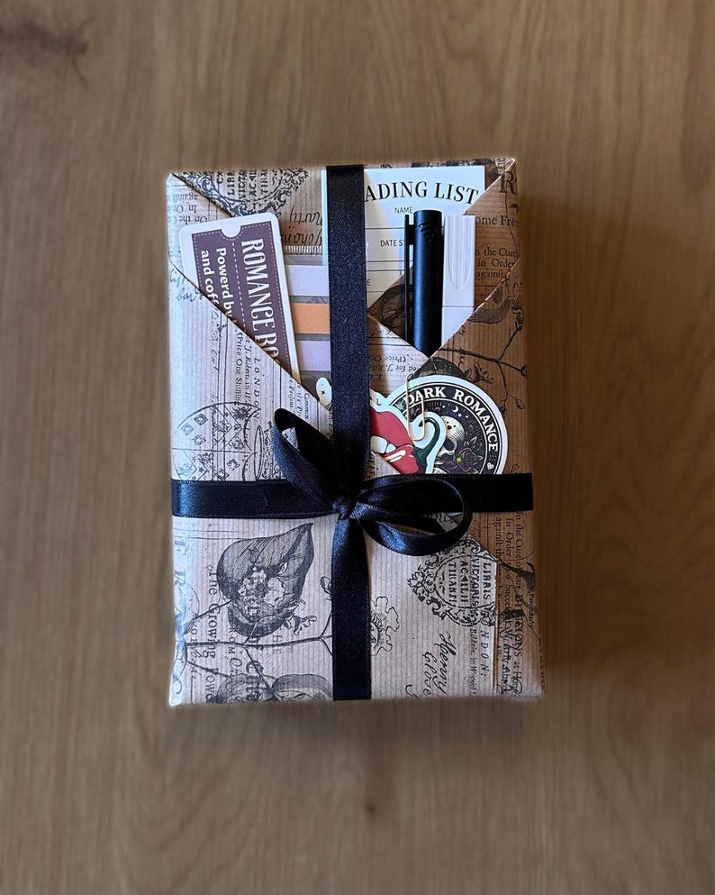 May include: A wrapped gift with a black ribbon and bow. The wrapping paper is brown with a vintage print. A pen, bookmark, and sticker are visible in a pocket on top. The bookmark reads "ROMANCE RO" and "Powered by and coffee".
