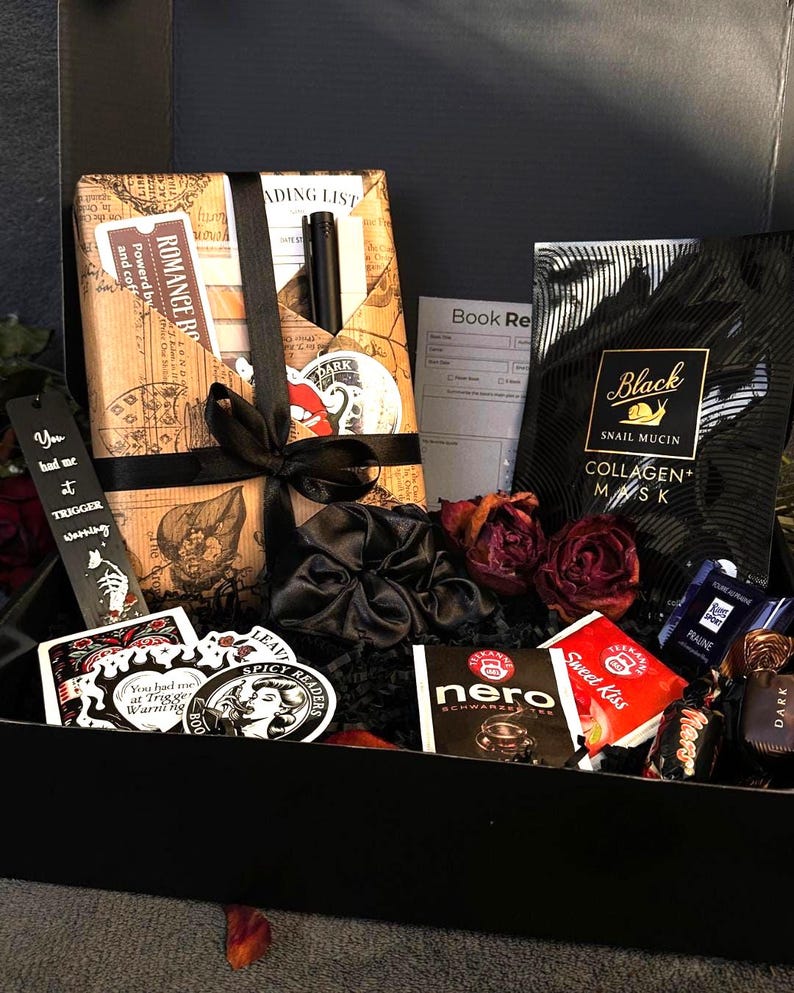 May include: A black gift box filled with various items. Includes a wrapped book, stickers, a bookmark, a black scrunchie, tea bags, a face mask, and chocolates. Dried red roses add a decorative touch.