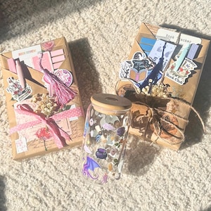 May include: A glass tumbler with a wooden lid, decorated with fantasy-themed stickers, sits between two wrapped packages. The packages are wrapped in brown paper and adorned with stickers, ribbons, and dried flowers. One package has the text "Book Review".
