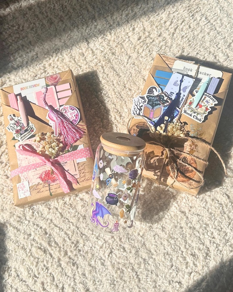 May include: A glass tumbler with a wooden lid, decorated with fantasy-themed stickers, sits between two wrapped packages. The packages are wrapped in brown paper and adorned with stickers, ribbons, and dried flowers. One package has the text "Book Review".