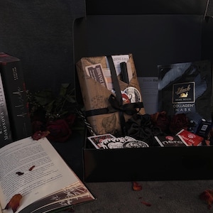 May include: A black gift box filled with various items, including books, a face mask, stickers, and wrapped packages tied with black ribbons. The open book displays text and fallen leaves.