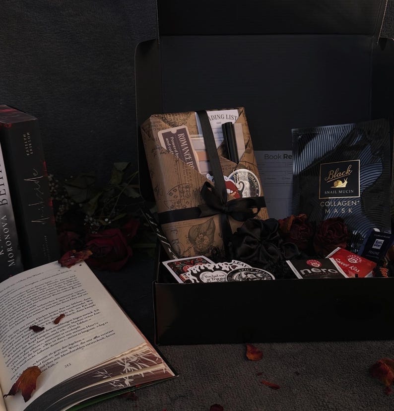 May include: A black gift box filled with various items, including books, a face mask, stickers, and wrapped packages tied with black ribbons. The open book displays text and fallen leaves.