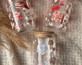 May include: Three clear glass tumblers with bamboo lids, each featuring colorful printed decals. The designs include stacks of books, roses, cowboy boots, hearts, ice skates, hockey jerseys, and various small decorative icons in pink, red, and blue.