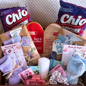 Könnte beinhalten: Eine Geschenkbox mit zwei Tüten Chio-Popcorn, zwei Gesichtsmasken, einer weißen Stumpenkerze, einem Paar flauschiger lavendelfarbener Socken, einem Paar hellblauer flauschiger Socken, einer rosa Haarspange, einem geblümten Haargummi, Teebeuteln und bedruckten Lese-Aktivitätskarten.