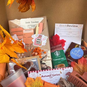 Könnte beinhalten: Eine Geschenkbox gefüllt mit herbstlichen Artikeln, darunter eine Buch-Tracker-Karte, ein Buchbewertungsformular, ein Starbucks-Cappuccino-Päckchen, Almar-Schokolade, eine Kerze, eine Haarklammer, ein Haargummi, Ritter Sport-Schokolade und dekorative Ahornblätter.