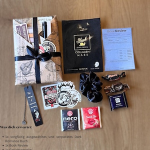 May include: A gift box with a wrapped book, face mask, scrunchie, bookmark, stickers, tea, and snacks. The book is wrapped in paper with a black ribbon. The face mask is black with white text. The bookmark has text.