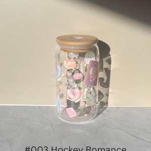 May include: A clear glass can-shaped tumbler with a wooden lid, decorated with pastel-colored illustrations of gifts, purses, and hearts. The text "#003 Hockey Romance" is printed at the bottom.