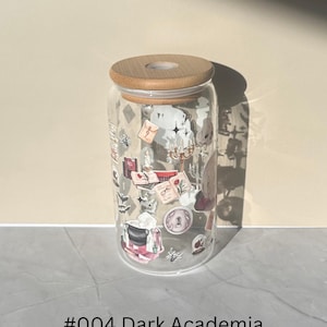 May include: A clear glass tumbler with a bamboo lid, decorated with stickers of books, candles, and other dark academia-themed elements. The text "#004 Dark Academia" is printed at the bottom. The tumbler is cylindrical in shape.