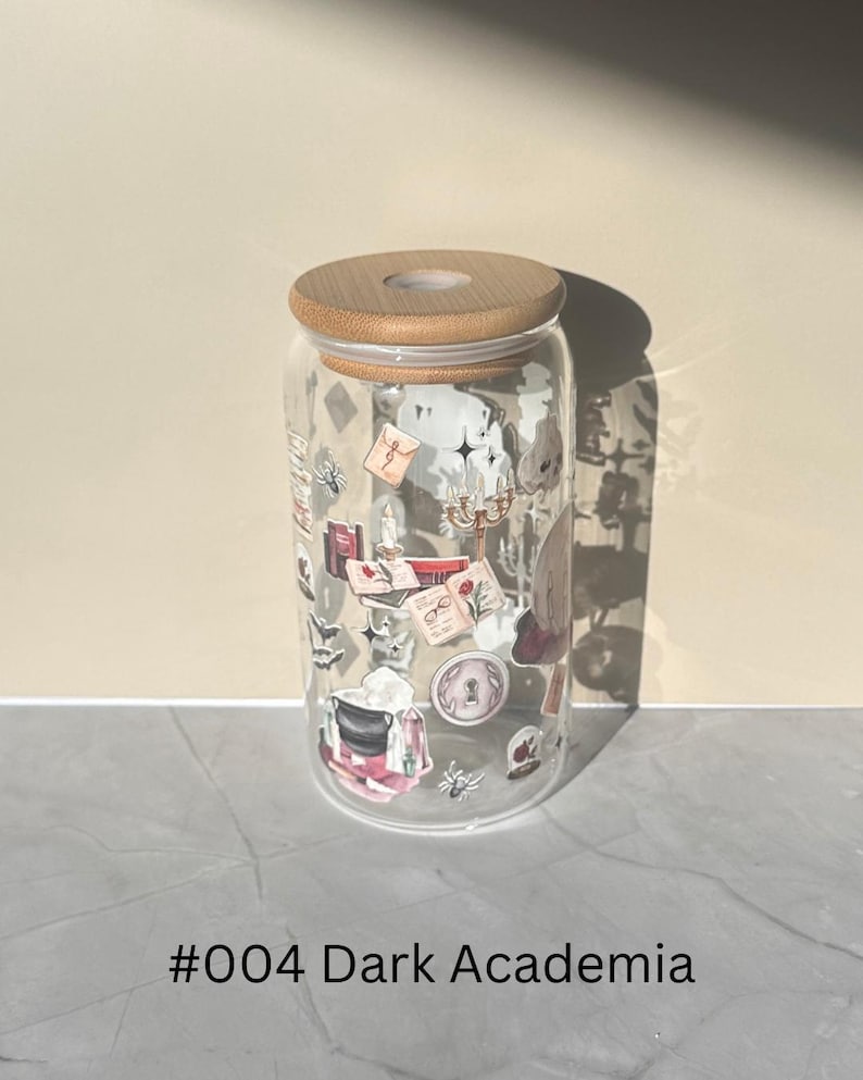 May include: A clear glass tumbler with a bamboo lid, decorated with stickers of books, candles, and other dark academia-themed elements. The text "#004 Dark Academia" is printed at the bottom. The tumbler is cylindrical in shape.