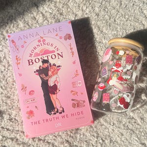 May include: A book titled "Mornings in Boston" by Anna Lane, with a pink cover featuring a couple embracing. Beside it is a clear glass jar with a wooden lid, decorated with stickers of hearts, flowers, and other small images.