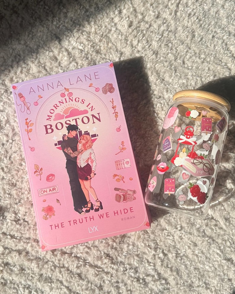 May include: A book titled "Mornings in Boston" by Anna Lane, with a pink cover featuring a couple embracing. Beside it is a clear glass jar with a wooden lid, decorated with stickers of hearts, flowers, and other small images.