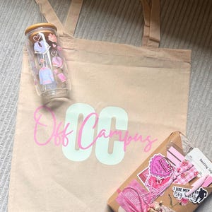 Könnte beinhalten: Eine beigefarbene Canvas-Tragetasche mit den Worten "Off Campus" in Rosa und Weiß. Auf der Tasche befindet sich ein Glasbecher mit Holzdeckel und Bekleidungsgrafiken. Ein verpacktes Geschenk mit rosa Akzenten und Aufklebern ist ebenfalls vorhanden.