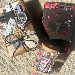 May include: A book, wrapped gift, and glass jar with a wooden lid. The book has a dark cover with floral and skull imagery. The jar is decorated with stickers, including a "Dark Book Club" design. The wrapped gift is tied with twine.