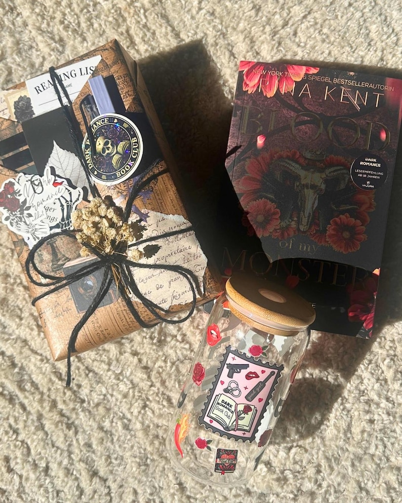 May include: A book, wrapped gift, and glass jar with a wooden lid. The book has a dark cover with floral and skull imagery. The jar is decorated with stickers, including a "Dark Book Club" design. The wrapped gift is tied with twine.