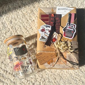 May include: A clear glass jar with a wooden lid and a wrapped package. The jar is covered in stickers with crime-themed graphics. The package is wrapped in brown paper and tied with twine, featuring stickers and a tassel. Text on the package reads "Plotting a Murder."