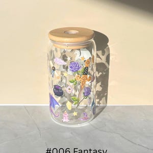 May include: A clear glass tumbler with a wooden lid, decorated with fantasy-themed illustrations. The design includes purple roses, a sword, a witch's hat, and a cauldron. The text "#006 Fantasy" is printed at the bottom.