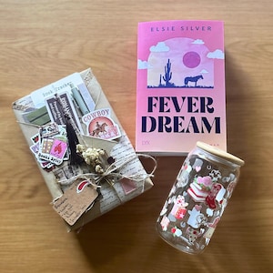 May include: An image featuring a wrapped gift, a pink book titled "Fever Dream", and a glass can with a wooden lid. The book cover displays a desert scene with a horse. The gift is adorned with stickers and a tag.