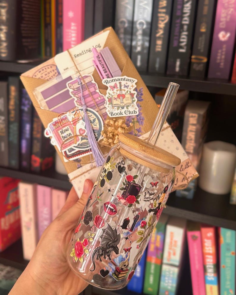 May include: A clear glass tumbler with a bamboo lid and straw, decorated with stickers of dragons, roses, and books. A wrapped package with stickers and the text "Reading Log" is held behind the tumbler. The background features a bookshelf filled with books.