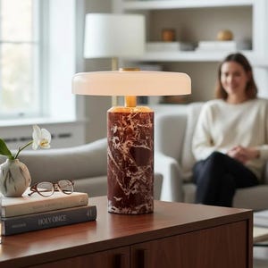Pode incluir: Uma moderna luminária de mesa com base cilíndrica com padrão de mármore vermelho e castanho. A luminária apresenta uma cúpula branca em forma de disco e um pescoço dourado. Está colocada sobre um armário de madeira, juntamente com livros, um copo e um pequeno vaso.