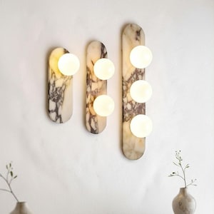 Può includere: Tre applique da parete in marmo con luci sferiche. Le applique sono orientate verticalmente, con una parte superiore e inferiore arrotondata. Il marmo presenta una miscela di venature beige, marroni e bianche. Le luci sono bianche.