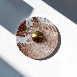 Puede incluir: Un posavasos redondo de mármol con un patrón marrón rojizo y blanco. Un pequeño pomo circular de latón está centrado en el posavasos. El posavasos está sobre una superficie blanca, con sombras.