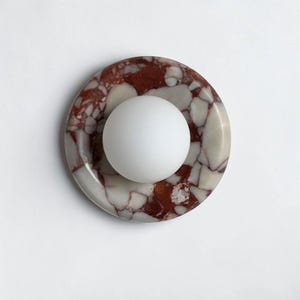 Puede incluir: Un objeto decorativo redondo con una esfera blanca centrada en una base de mármol moteado, de color marrón rojizo y crema. El mármol tiene un patrón veteado natural. El objeto está sobre un fondo blanco liso.