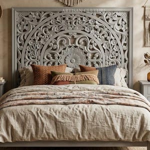 Könnte beinhalten: Ein grauer Bettrahmen aus Holz mit einem großen, kunstvoll geschnitzten Kopfteil mit Blumen- und Mandalamustern. Das Bett ist mit einer beigen Leinenbettdecke und mehreren dekorativen Kissen in Orange-, Braun- und Blautönen bezogen.