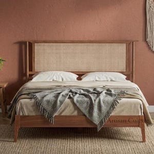 Könnte beinhalten: Ein Holzbettgestell mit einem geflochtenen Rohrgeflecht-Kopfteil, ausgestattet mit weißen Kissen und einer grauen, strukturierten Tagesdecke. Das Bett steht in einem Raum mit terrakottafarbener Wand, einer hängenden Korblampe und einem Makramee-Wandbehang.