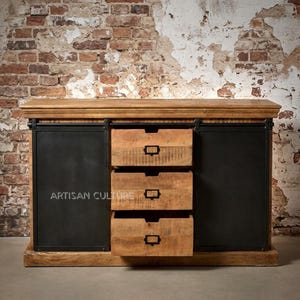 Puede incluir: Gabinete de madera con tres cajones y dos puertas correderas de metal negro. El gabinete tiene un acabado de madera natural con vetas visibles y el texto "ARTISAN CULTURE". El gabinete está contra una pared de ladrillo.