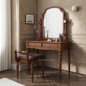 Puede incluir: Un tocador de madera con un gran espejo arqueado, dos cajones con tiradores de metal ornamentados y patas torneadas. Un taburete a juego con asiento tapizado en marrón oscuro y respaldo curvo está colocado frente al tocador.